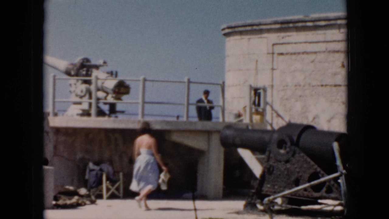 1956 Cuba home-movie frame showing a woman in a pale dress near a large cannon, with a suited man on a raised platform behind her.