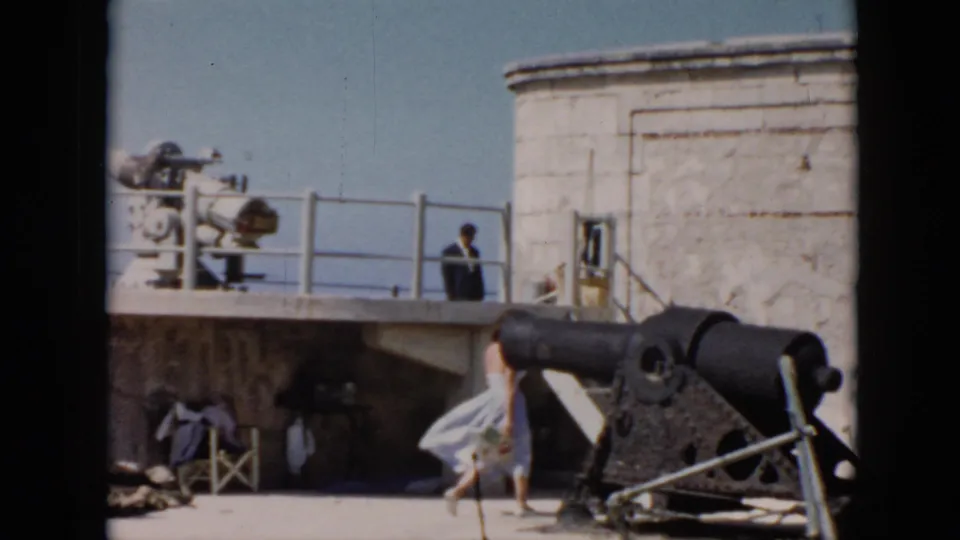 1956 Cuba frame of a woman moving past a dark cannon at a historic fort, with stonework, railing, and another visitor visible behind her.