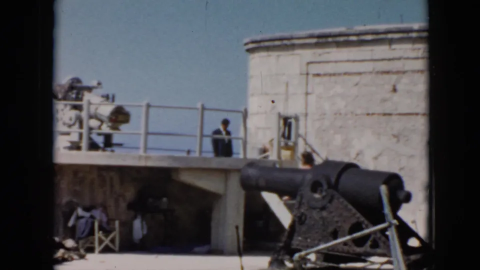 1956 Cuba home-movie frame with the cannon dominating the foreground and a visitor partly visible near the stone fort wall and railing.