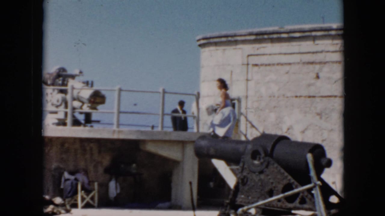 1956 Cuba home-movie frame showing a black cannon, whitewashed fort walls, railings, visitors, and blue sky near the coast.