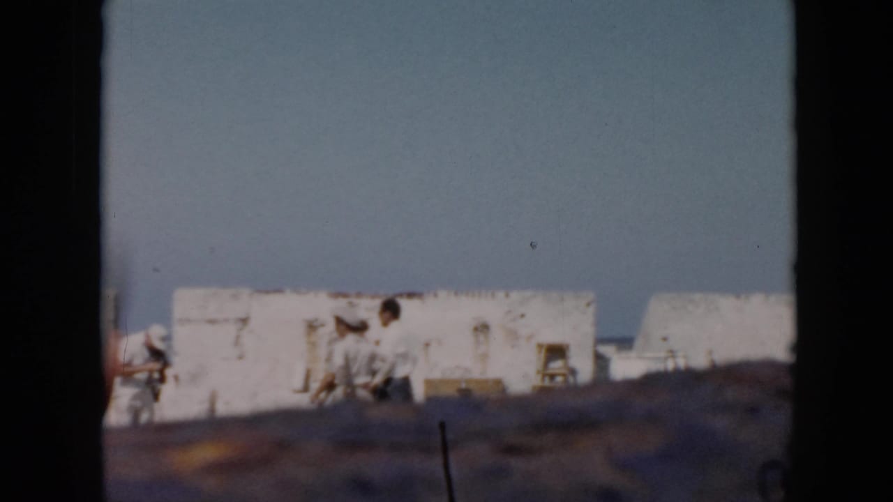 1956 Cuba frame with distant visitors beside white fort walls and the sea, partly blocked by the camera position or film edge.