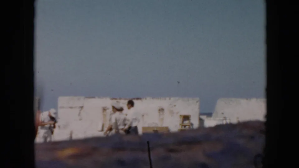 1956 Cuba frame with distant visitors beside white fort walls and the sea, partly blocked by the camera position or film edge.