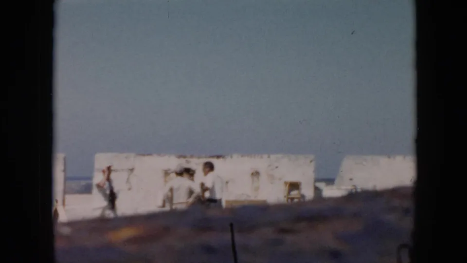 1956 Cuba home-movie frame showing visitors along white fort walls, with open sky, sea, and blurred foreground framing.