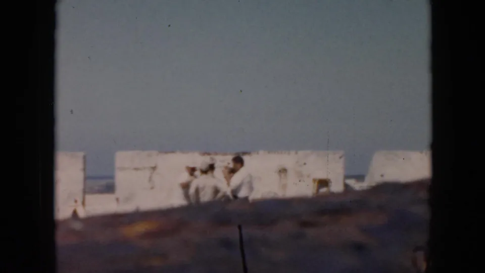 1956 Cuba home-movie frame showing tourists beside weathered white fortress walls with sea and sky beyond.