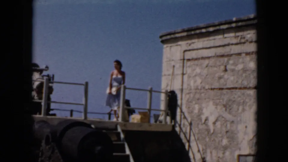 Woman in a light dress walks on a raised fortress platform near railings and a cannon-like object in Cuba.