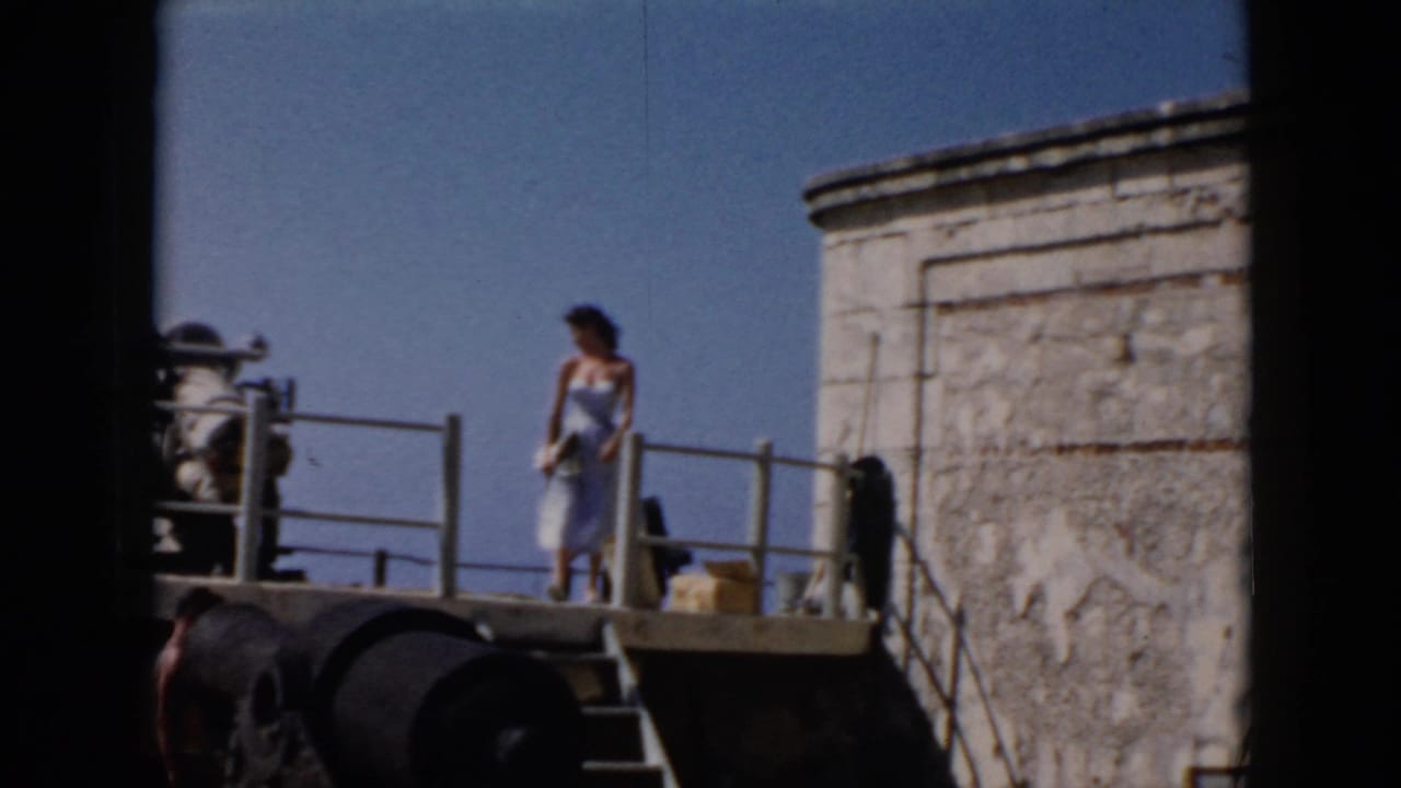 In 1956 Cuba footage, a woman in a blue dress poses on a stone fort platform with railings, cannon-like forms, and blue sky behind her.