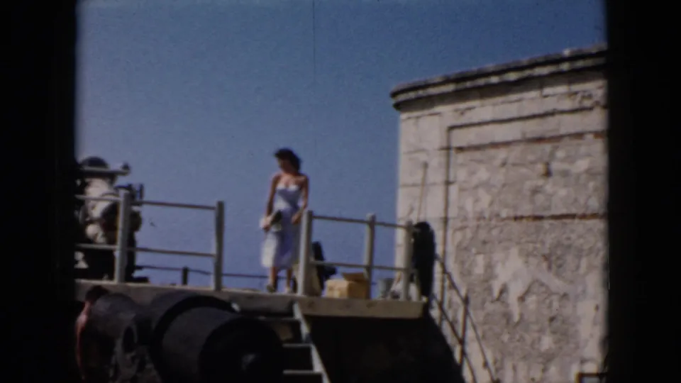 In 1956 Cuba footage, a woman in a blue dress poses on a stone fort platform with railings, cannon-like forms, and blue sky behind her.