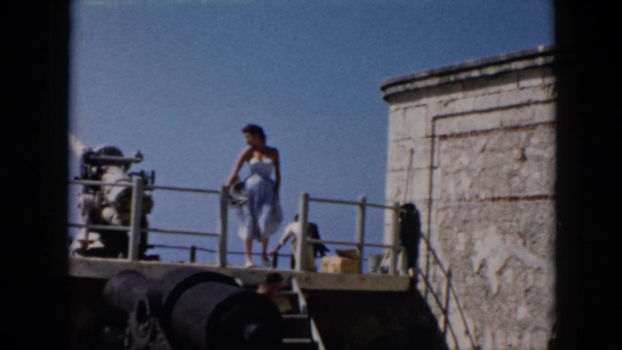 In 1956 Cuba footage, the woman turns on the fort platform near railings, stonework, cannon-like objects, and a small yellow container.
