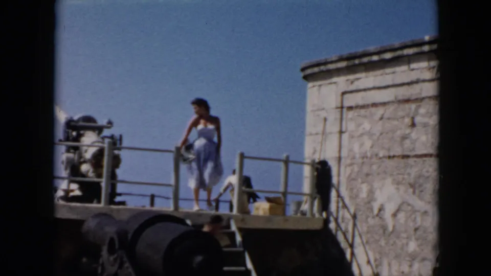 In 1956 Cuba footage, the woman turns on the fort platform near railings, stonework, cannon-like objects, and a small yellow container.