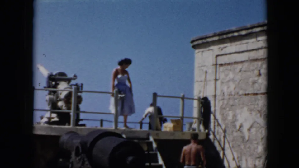 In 1956 Cuba footage, the woman stands on the fort platform while a shirtless man is visible below, with railings, stonework, and cannon-like forms nearby.