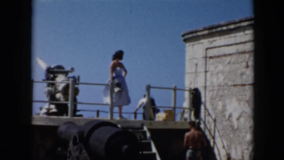 In 1956 Cuba footage, the woman faces across the fort platform with railings and stonework visible as a shirtless man stands below at right.