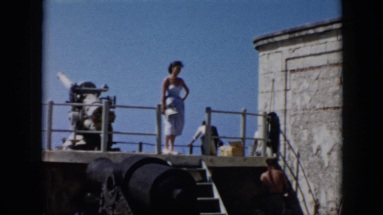 1956 Cuba home-movie frame showing a woman in a light blue dress standing near a cannon on a stone fort platform.