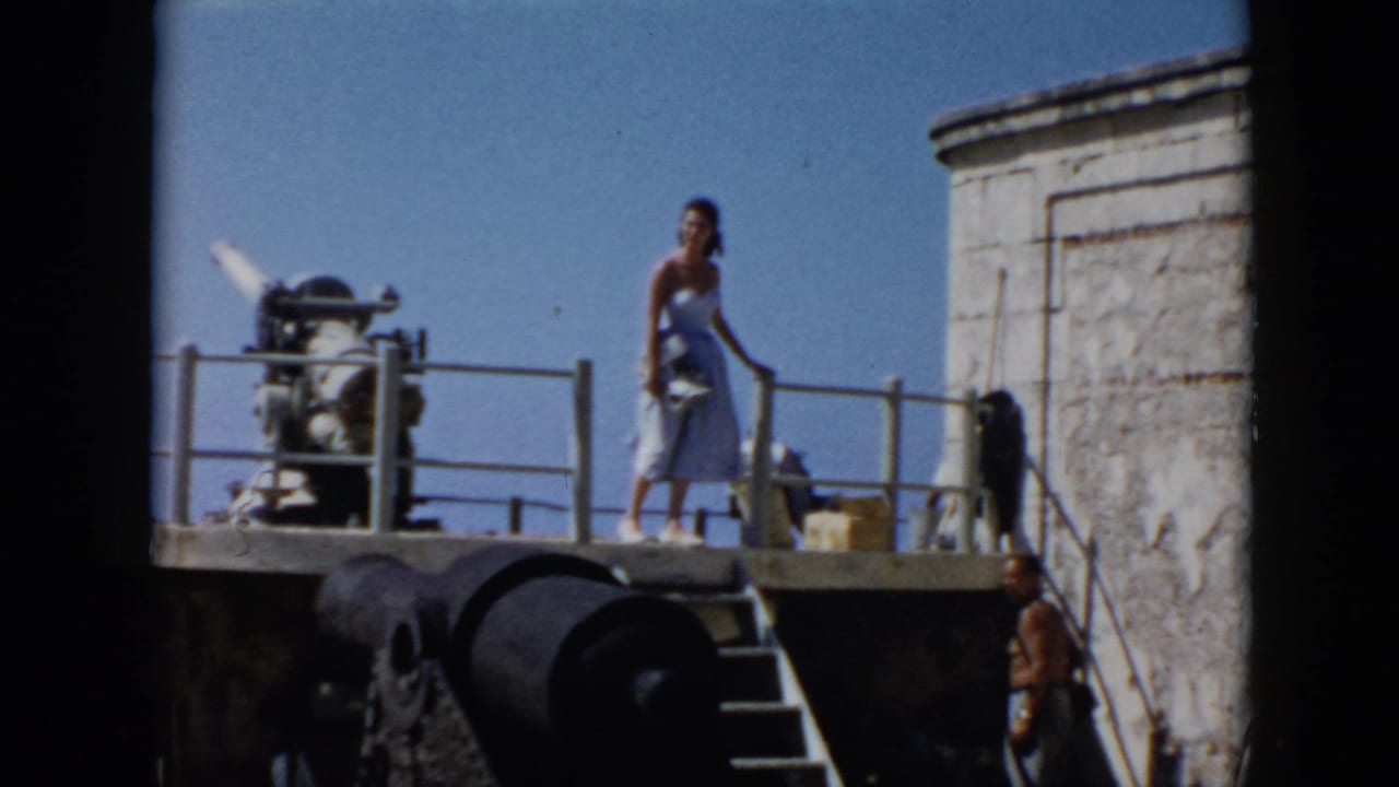 1956 Cuba frame with a woman standing on a fort platform beside a cannon, with another visitor near the steps.
