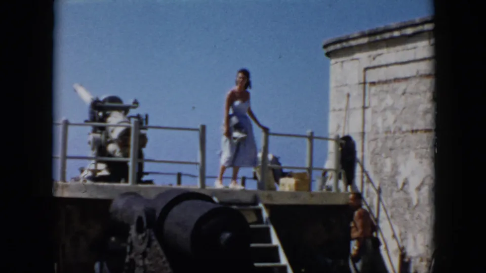 1956 Cuba frame with a woman standing on a fort platform beside a cannon, with another visitor near the steps.