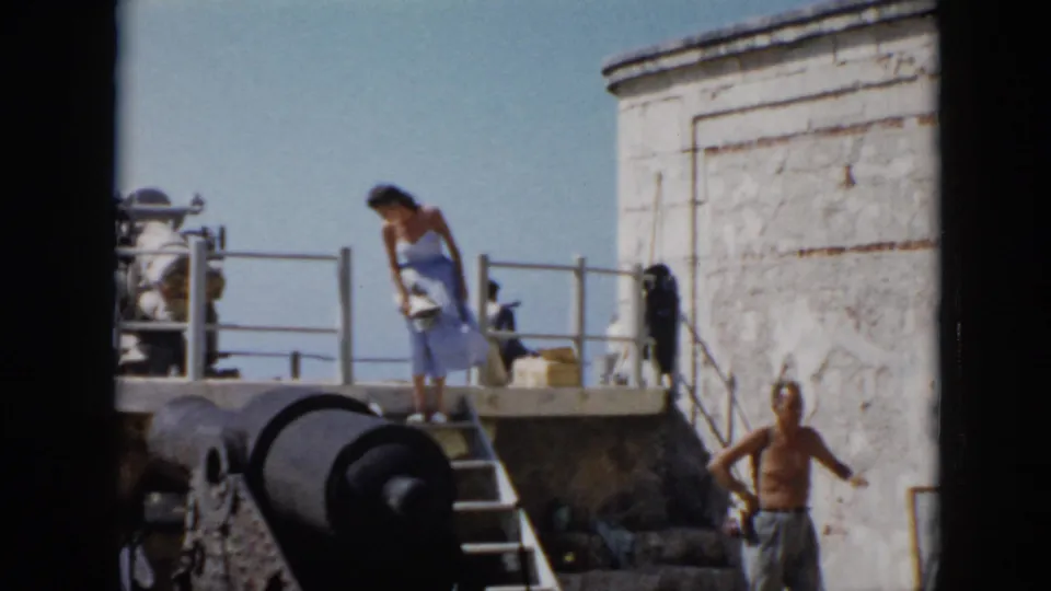 1956 Cuba home-movie frame showing a woman near fort steps and cannon, with a shirtless man visible below.
