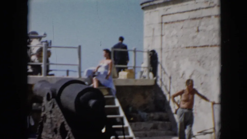 1956 Cuba frame showing a woman seated beside a cannon at a fort, with other visitors on the platform.