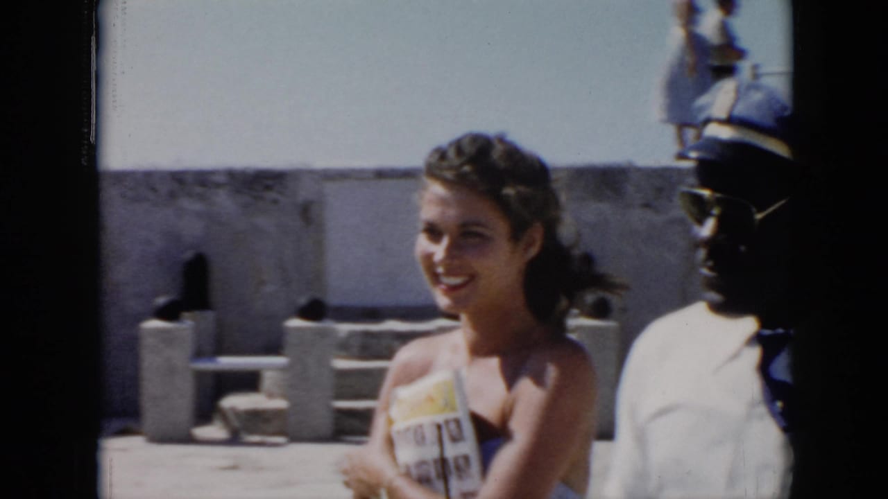 1956 Cuba frame of a smiling woman holding a magazine near an apparent uniformed official in a sunlit stone fort courtyard.