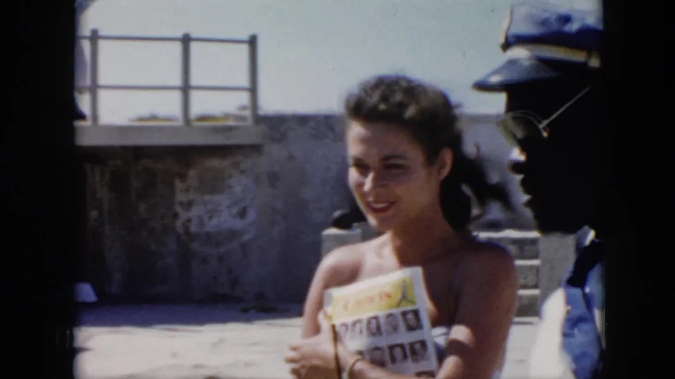 1956 Cuba home-movie frame with a woman holding a magazine as a uniformed man stands partly cropped at the edge of the courtyard scene.