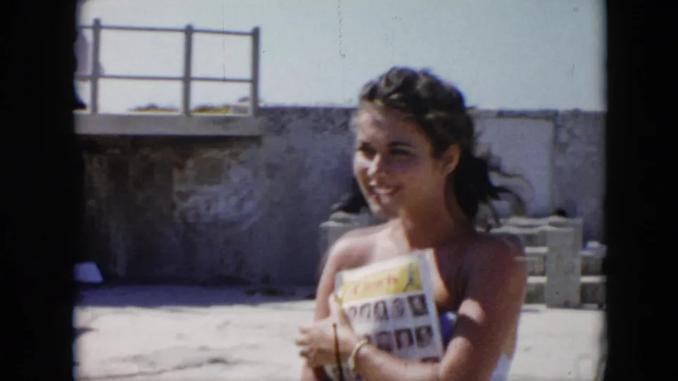 1956 Cuba frame showing a smiling woman with a magazine in a bright stone fortress courtyard with wall and railing behind her.