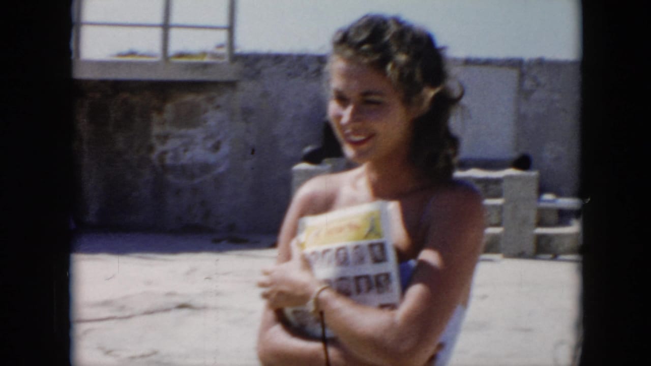In Cuba home-movie footage from 1956, a young woman smiles in bright sun while holding a printed magazine near concrete walls.