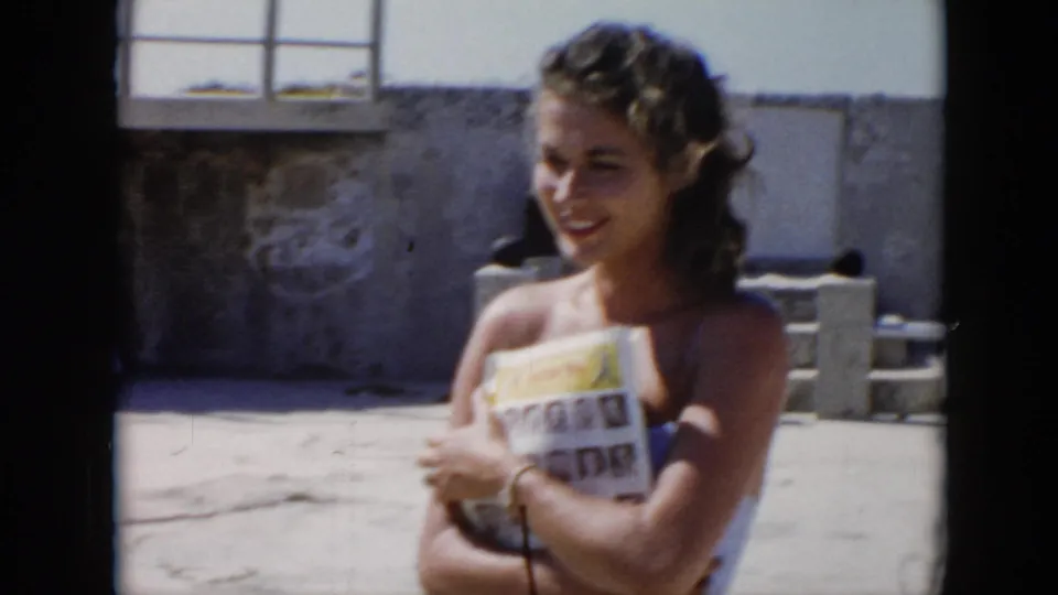 In Cuba home-movie footage from 1956, a young woman smiles in bright sun while holding a printed magazine near concrete walls.