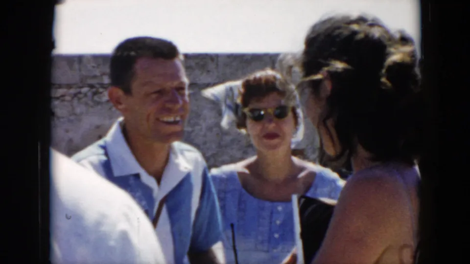 In a 1956 Cuba outdoor scene, a man in a light blue shirt and a woman in sunglasses face a woman seen from behind.