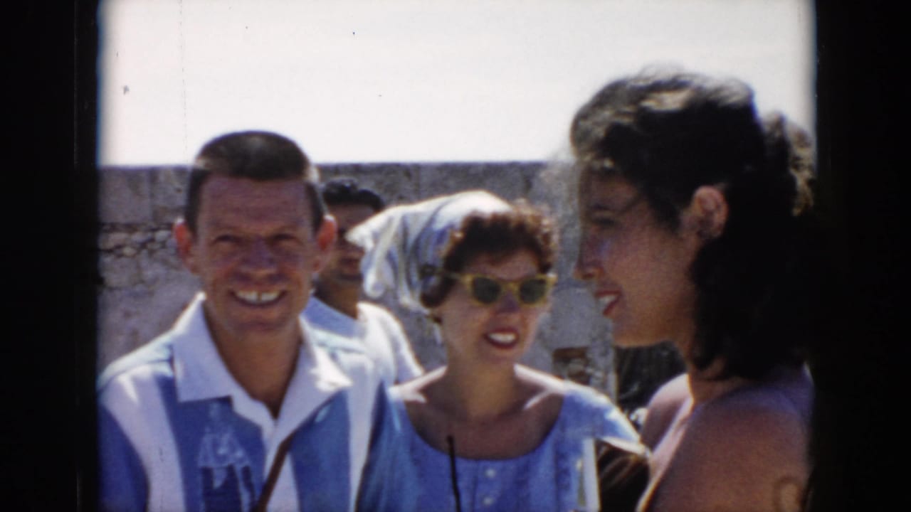 In 1956 Cuba, adult tourists stand close together near a bright stone wall, with one man smiling toward the camera.