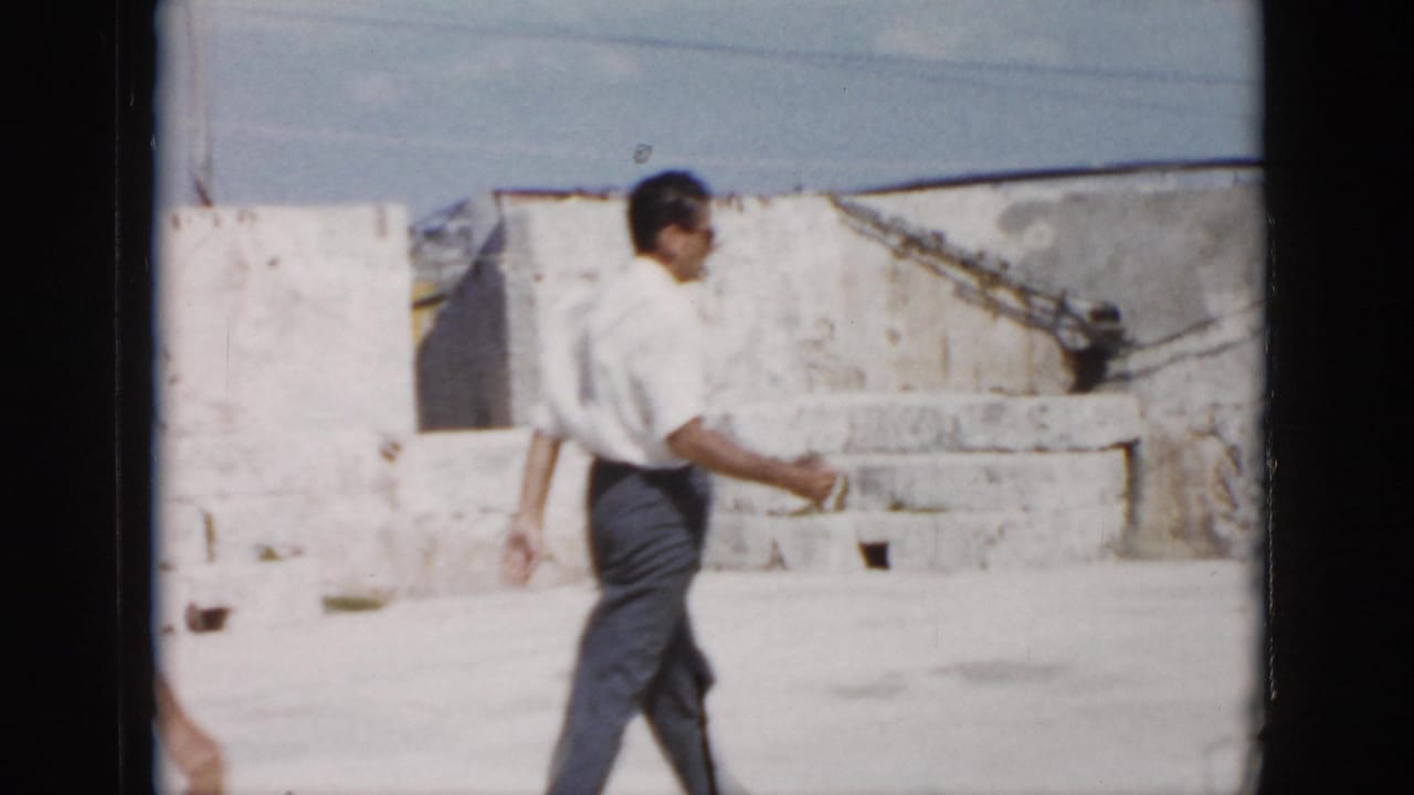 An adult man in a white shirt and dark trousers walks through a sunlit stone courtyard in Cuba.