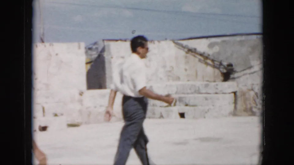 An adult man in a white shirt and dark trousers walks through a sunlit stone courtyard in Cuba.