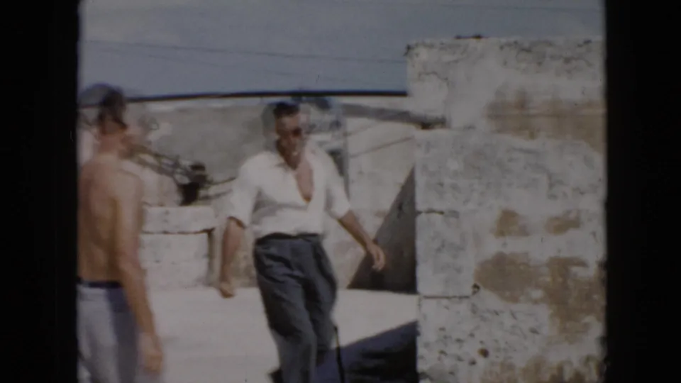 Two adult men move through a sunlit ruin courtyard, one in a white shirt and another partly shirtless at frame edge.