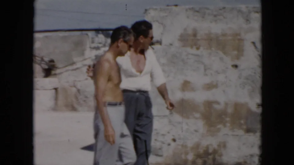 Two adult men walk together beside a weathered wall in the Cuba ruin setting, one shirtless and one in a white shirt.
