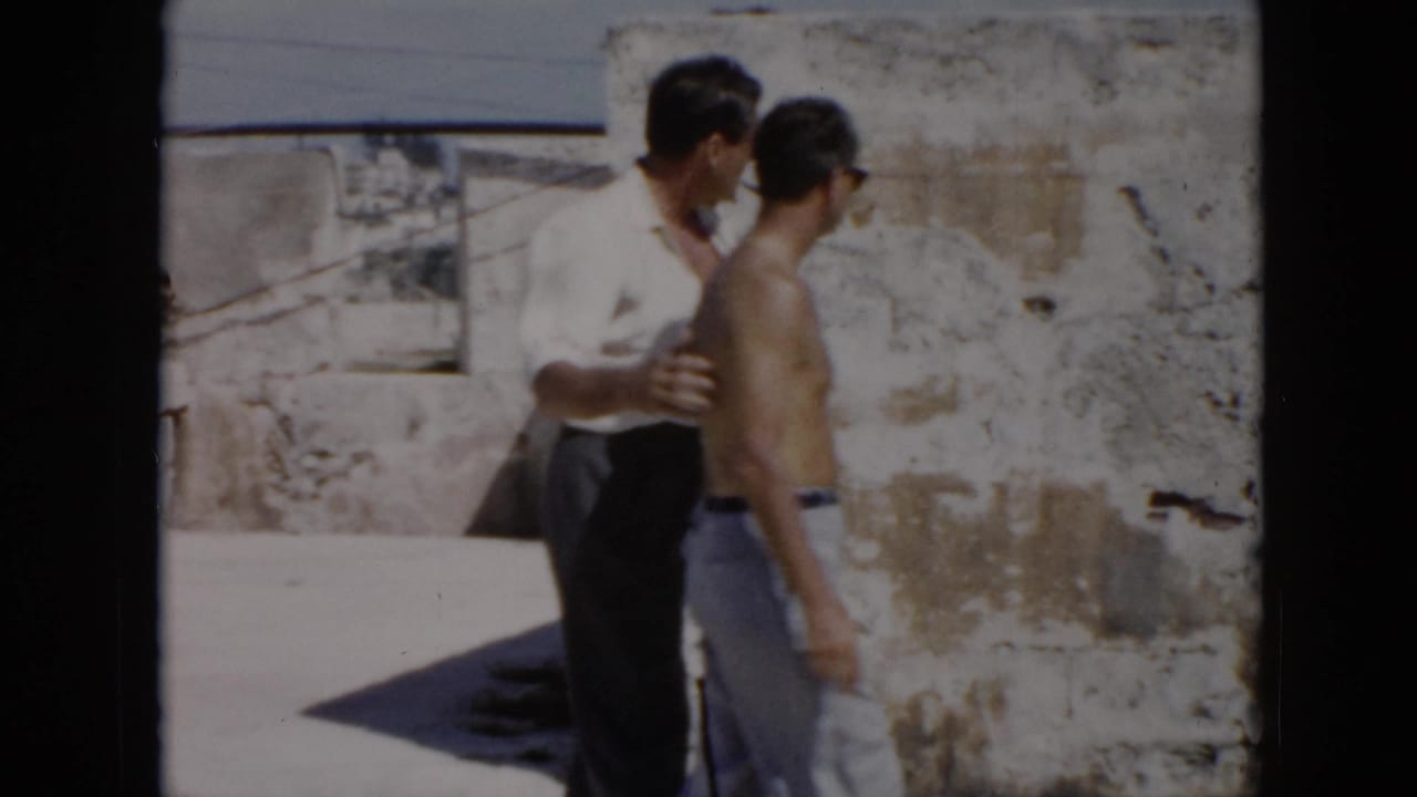 In 1956 Cuba footage, a clothed man appears to guide a shirtless man beside sunlit stone walls.