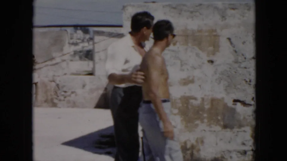 In 1956 Cuba footage, a clothed man appears to guide a shirtless man beside sunlit stone walls.