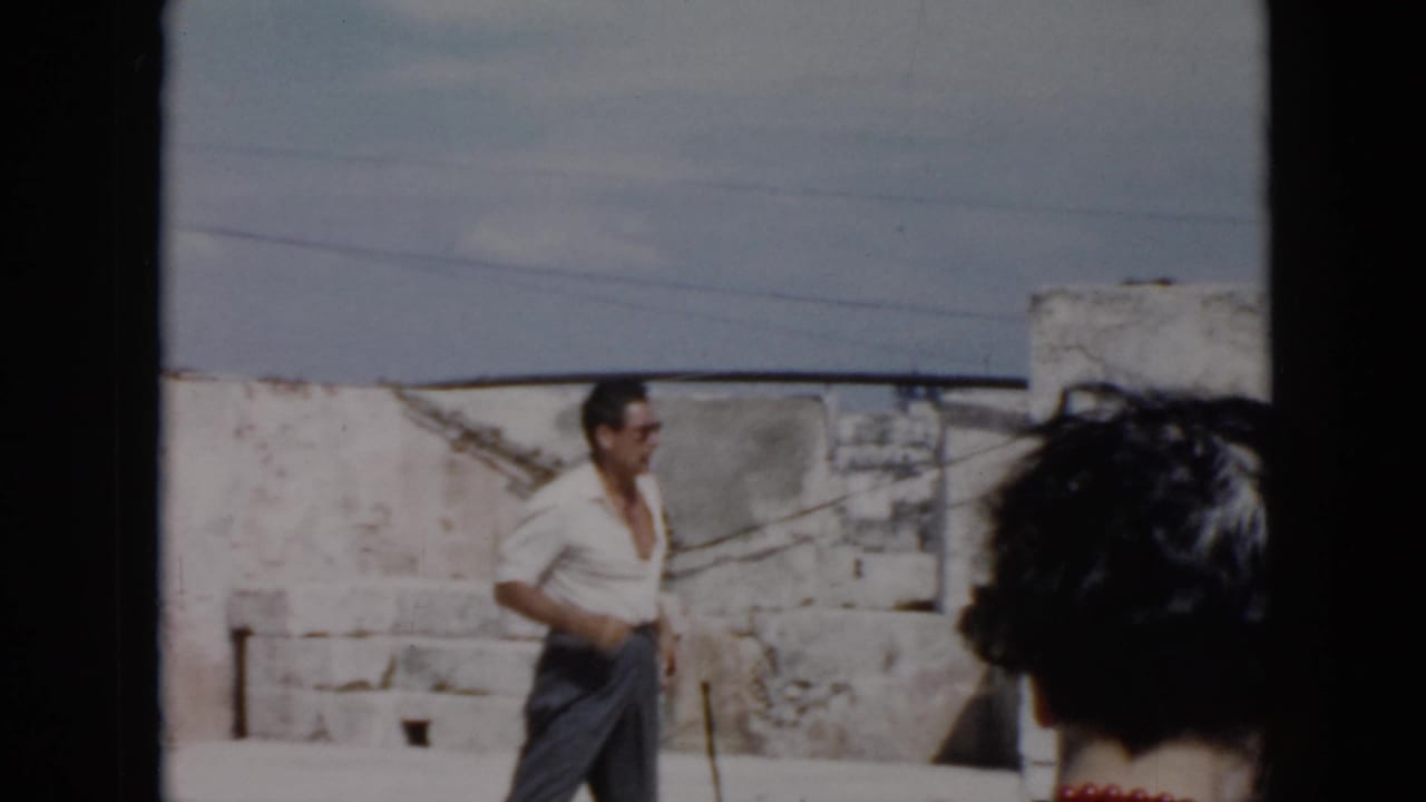 Man in a white shirt walks near stone walls while another person is seen from behind.
