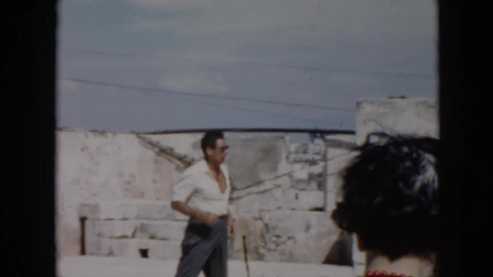 A man in a white open-collar shirt walks across a sunlit stone-walled area in Cuba, with another figure in the foreground.