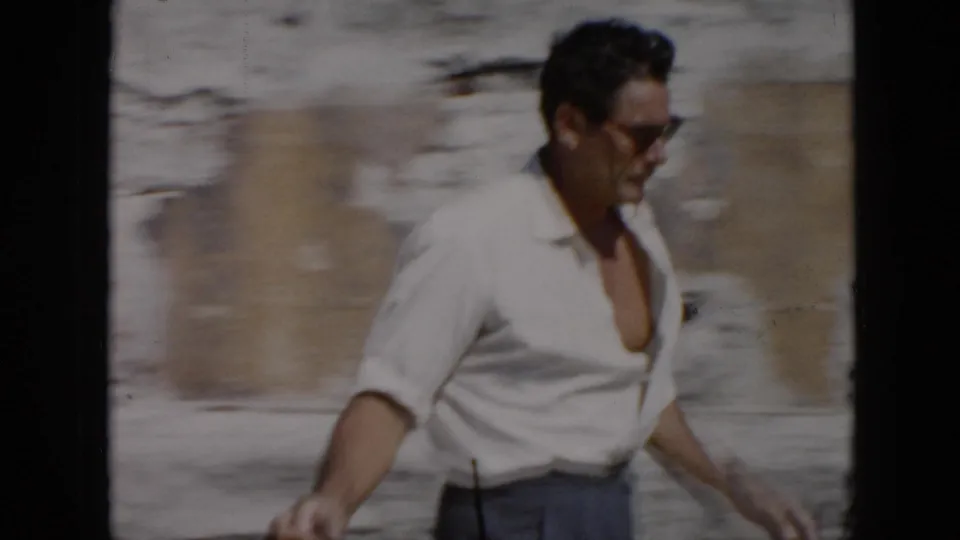 A man in sunglasses and an open-collar white shirt passes close to the camera in sunlit 1956 Cuba footage.