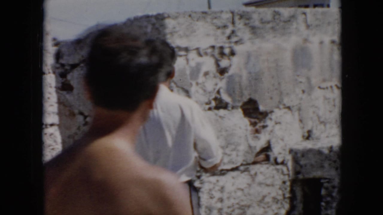 Shirtless man seen from behind while another man stands near rough stone blocks.