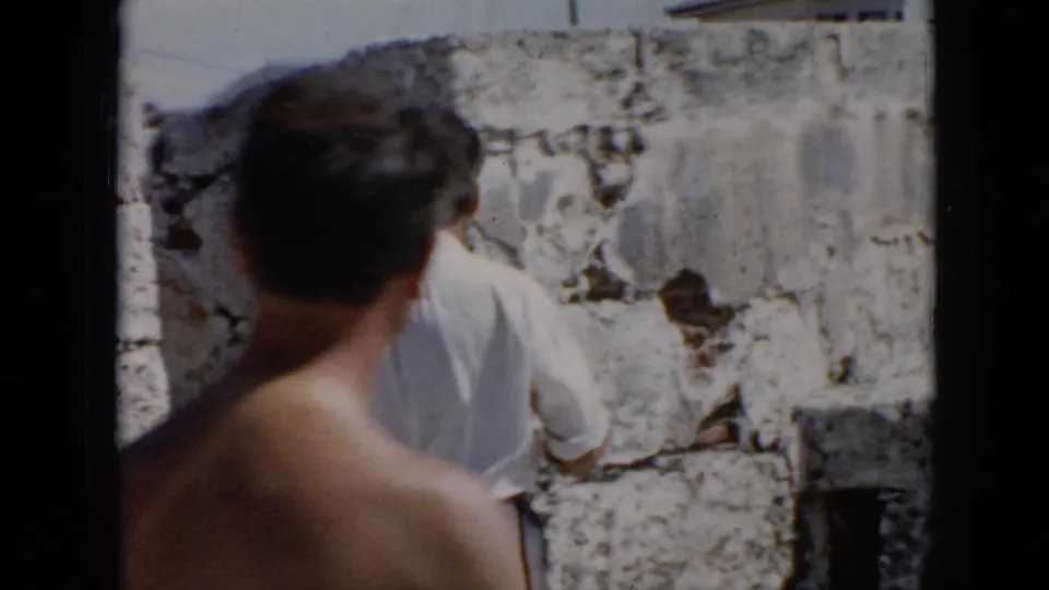 1956 Cuba travel footage frame with a shirtless man in the foreground and another visitor near worn stone masonry.