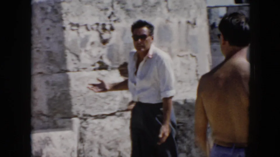 1956 Cuba frame showing a man in sunglasses gesturing beside weathered stone blocks as another visitor stands nearby.
