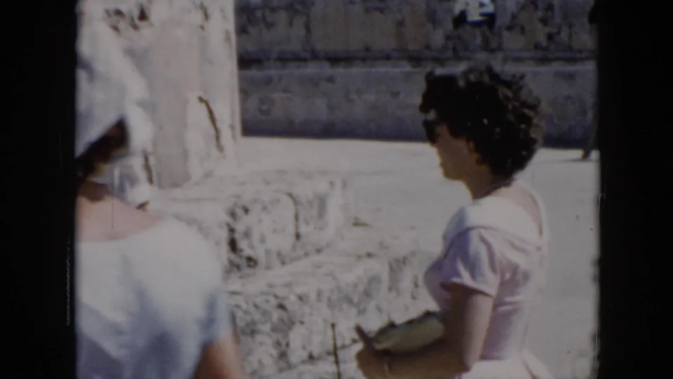 1956 Cuba home-movie frame showing a woman in a light dress and hat walking past sunlit stone ruins.
