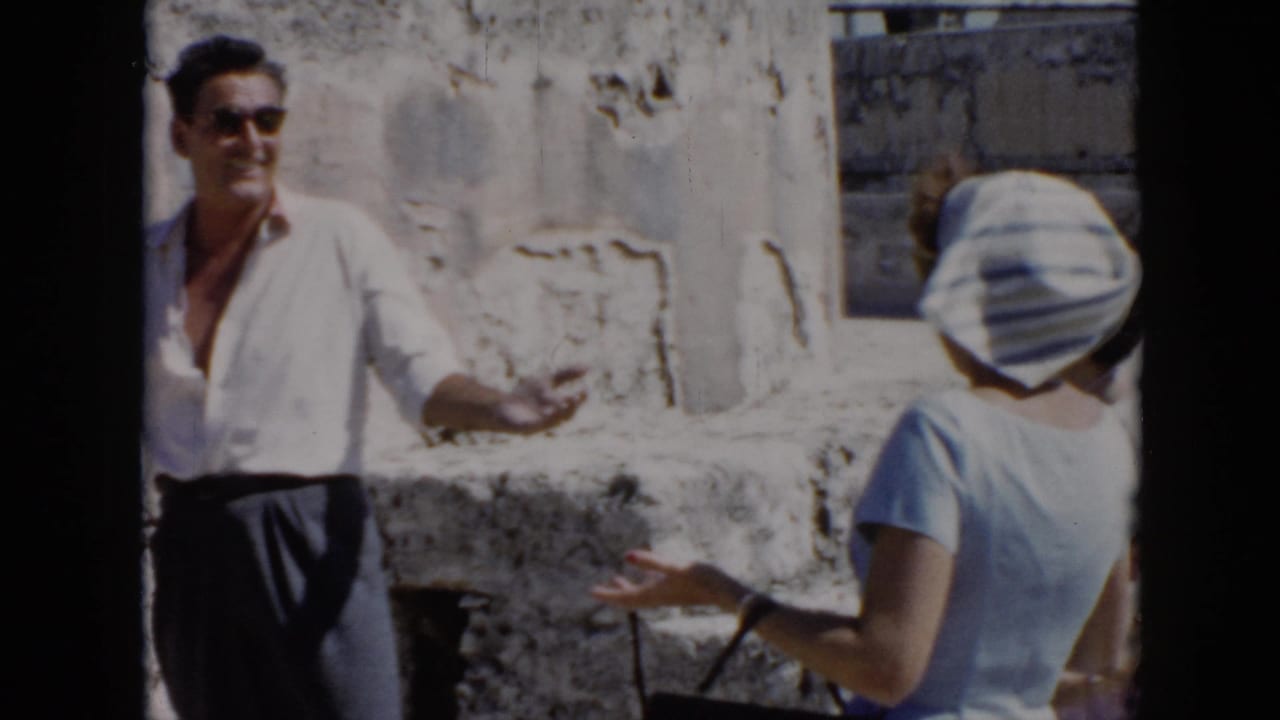 1956 Cuba frame with a man in a white shirt and sunglasses gesturing while speaking to a woman in a striped headscarf near stone ruins.