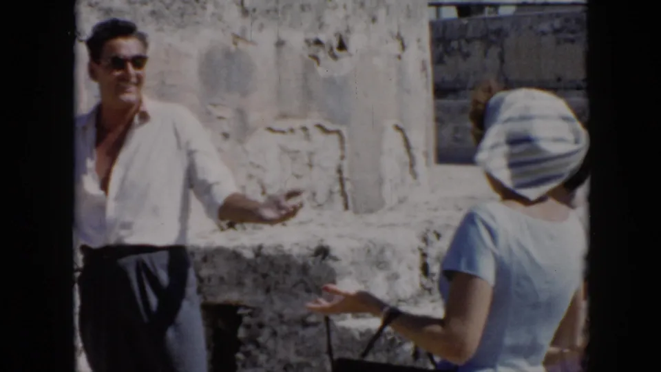 1956 Cuba frame with a man in a white shirt and sunglasses gesturing while speaking to a woman in a striped headscarf near stone ruins.
