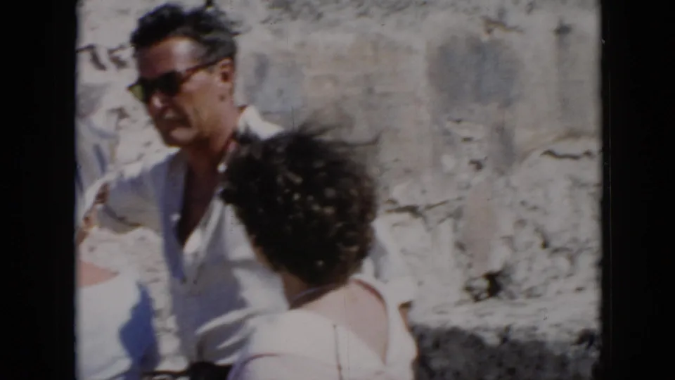 Close 1956 Cuba home-movie view of a man in sunglasses and two women conversing beside weathered stone masonry in bright sun.