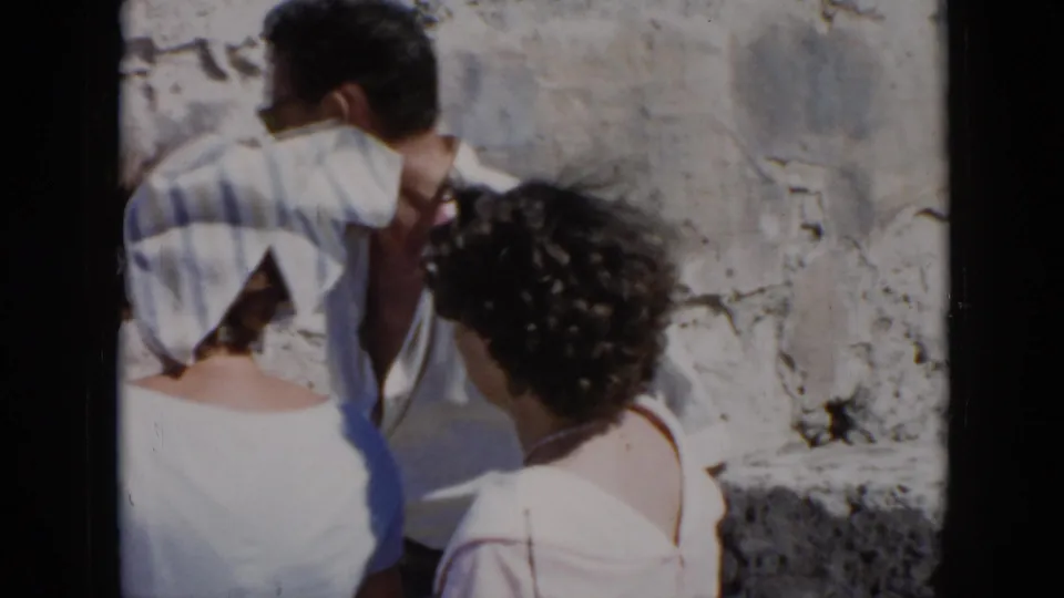 1956 Cuba travel-home-movie frame showing two women and a man turned toward each other beside weathered stonework, with faces partly obscured.
