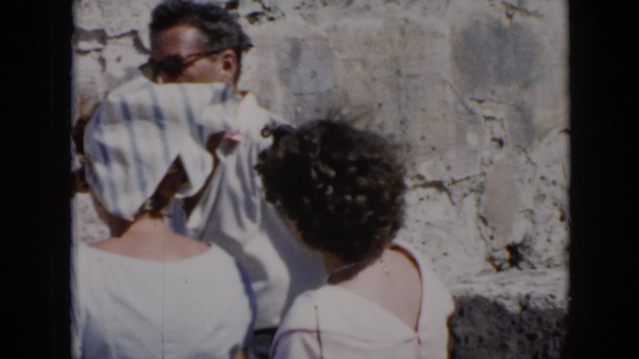 In 1956 Cuba home-movie footage, adults in light summer clothing pass a weathered stone wall at a historic outdoor site.