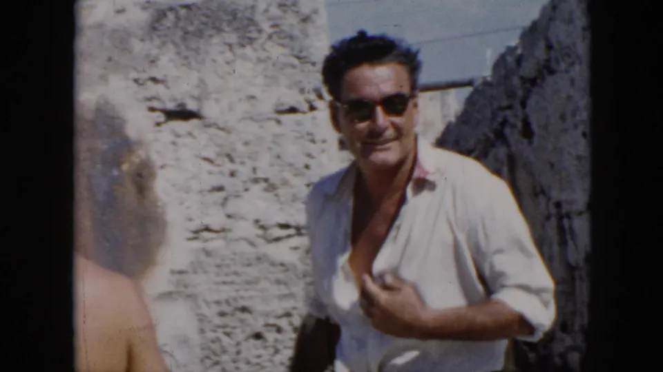 A man in a white shirt and sunglasses smiles toward the camera beside weathered stonework in 1956 Cuba footage.