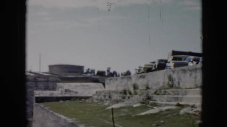 The camera looks across an open stone site in Cuba toward parked 1950s cars and a distant fort-like structure.
