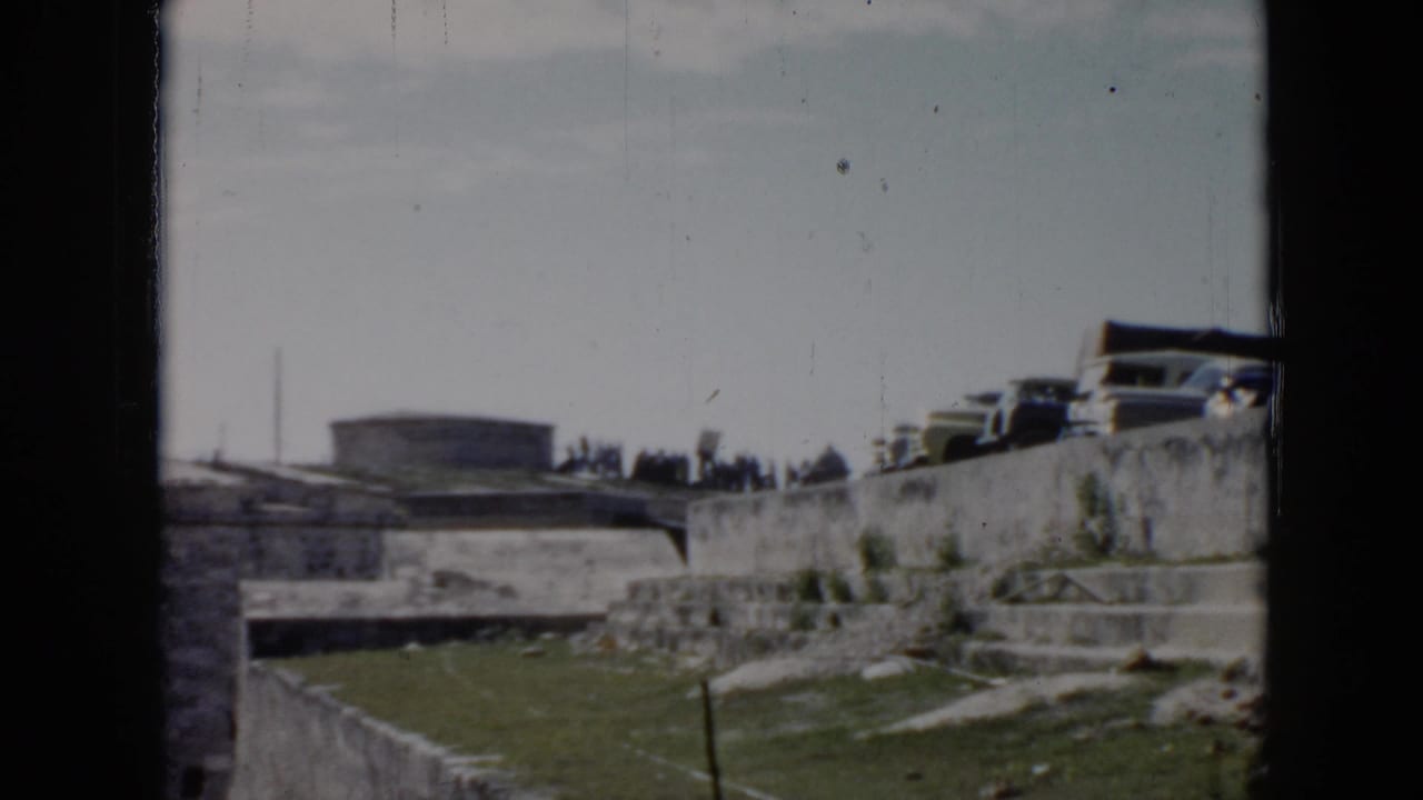 1956 Cuba 8mm frame showing stone walls, steps, grass, parked cars, and distant people under a bright sky.