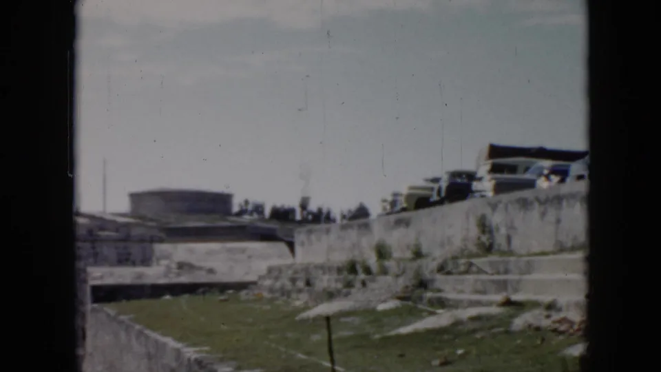 1956 Cuba 8mm frame with parked cars on an elevated stone-walled area, grass below, and distant people near the horizon.