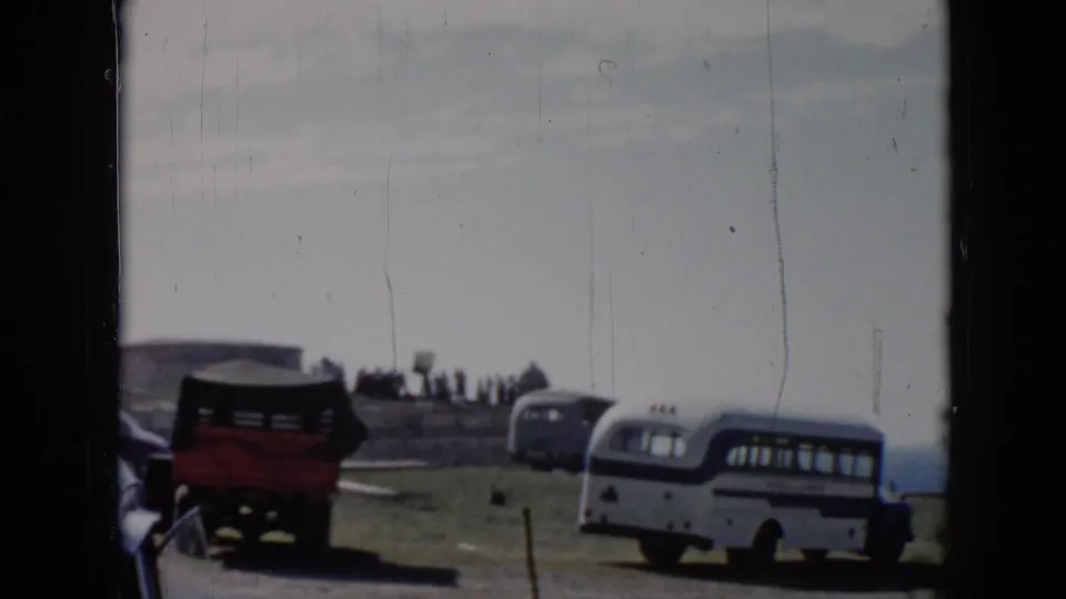 1956 Cuba 8mm frame showing a white bus, red truck, parked cars, stonework, and distant people in an outdoor parking area.